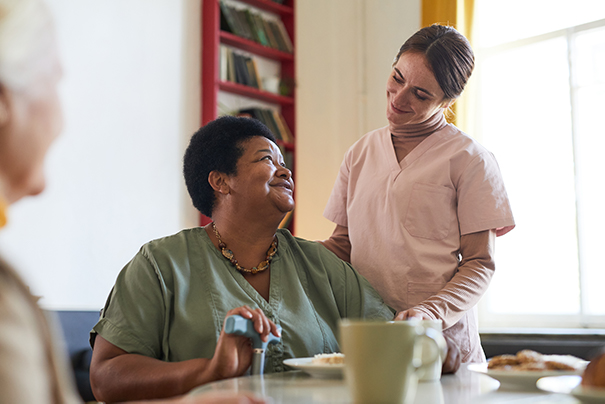 A caregiver in scrubs interacts warmly with a smiling older woman at a dining table, suggesting a supportive elderly care environment.