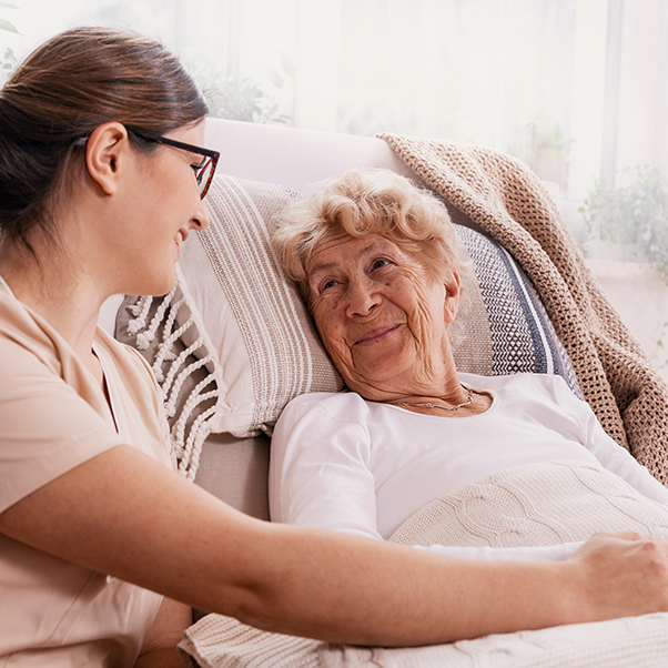 A caregiver smiles and interacts with an elderly woman sitting on a couch, showcasing warmth and companionship in a cozy setting.