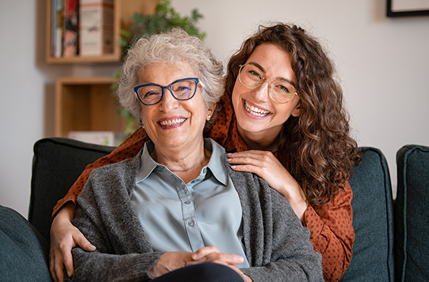 Two women sharing a joyful moment on a couch, one an older woman with curly gray hair and glasses, and the other a younger woman with long curly hair and glasses, both smiling warmly. The image conveys themes of family, love, and connection.